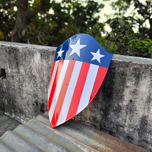 Puede incluir: Un objeto en forma de escudo con una parte superior azul con tres estrellas blancas y una parte inferior a rayas rojas y blancas. El escudo descansa sobre una superficie de metal corrugado, con un fondo verde borroso de follaje y una pared de hormig&oacute;n.