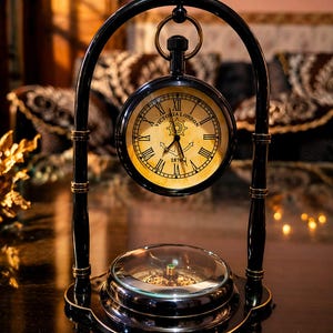 Puede incluir: Un reloj de estilo vintage negro y dorado con el texto "Victoria London" en la esfera. El reloj está sostenido por un soporte de metal negro con una brújula en la base. El reloj tiene números romanos y una esfera dorada.