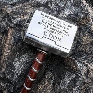 Può includere: Un martello in metallo argentato con l'iscrizione "Whosoever holds this hammer, if he be worthy, shall possess the power of... Thor." L'impugnatura è avvolta in pelle marrone con dettagli argentati. Il martello poggia su una superficie rocciosa grigia.