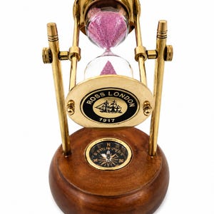 May include: A brass hourglass with pink sand, a compass, and a wooden base. The hourglass has a clear glass chamber and a brass frame. The base has a compass and a plaque with the text "ROSS LONDON 1917" and a ship illustration.