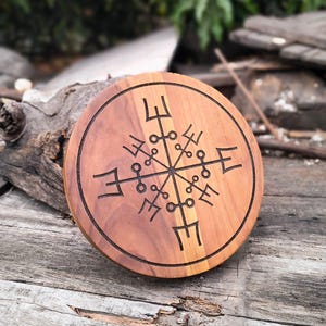 Könnte beinhalten: Runder Holzuntersetzer mit einem geschnitzten Vegvisir-Symbol. Der Untersetzer besteht aus hellem und dunklem braunem Holz und hat ein dunkelbraunes eingraviertes Design. Er liegt auf einer verwitterten Holzoberfläche.