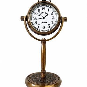 May include: A vintage-style brass desk clock with a white clock face and black numerals. The clock has a quartz movement and the word "Quartz" is printed on the face. The clock is supported by a decorative stand with an ornate base.