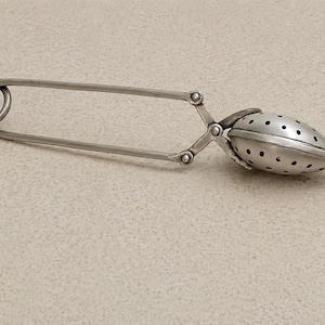 Op de afbeelding: Een zilverkleurige metalen thee-ei in de vorm van een grote veiligheidsspeld. Het voorwerp heeft een lange, slanke steel met een ronde lus aan het ene uiteinde en een geperforeerde, amandelvormige zeef aan het andere uiteinde voor losse thee.