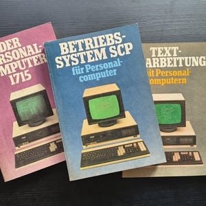 Może przedstawiać: Trzy zabytkowe książki techniczne z ilustracjami komputerów na okładkach. Lewa książka jest w kolorze magenty z napisem DER PERSONAL-COMPUTER 1715. Środkowa książka jest niebieska z napisem BETRIEBS-SYSTEM SCP. Prawa książka jest szara z napisem TEXT-ARBEITUNG.