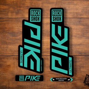 Könnte beinhalten: Zwei vertikale schwarze Aufkleber mit türkisfarbener Schrift, die das RockShox-Logo und das Wort PIKE ULTIMATE zeigen. Darunter befinden sich zwei kleinere horizontale Aufkleber, einer mit der Aufschrift RockShox PIKE und der andere mit RAPID RECOVERY REBOUND und Symbolen.