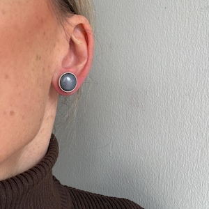 Peut inclure: Gros plan d'une oreille portant une boucle d'oreille grise. La boucle d'oreille est une pierre ronde grise avec un bord argenté. La personne porte un pull à col roulé marron.