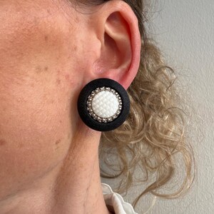 Puede incluir: Pendientes redondos de botón en blanco y negro. Los pendientes tienen un borde negro, un centro blanco con un patrón texturizado y un ribete plateado. Se muestran puestos en una oreja.
