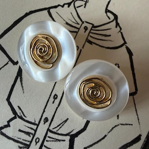 Puede incluir: Par de pendientes vintage. Cada pendiente presenta una base redonda blanca nacarada con un diseño de espiral dorado en el centro. Los pendientes están colocados sobre un fondo de ilustración vintage.