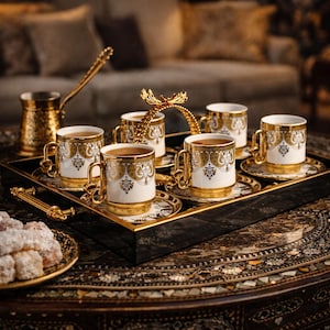 Könnte beinhalten: Ein Set aus sechs weiß-goldenen Kaffeetassen mit Untertassen, angeordnet auf einem schwarzen Tablett mit goldenen Griffen. Die Tassen weisen aufwendige goldene Details auf und sind mit Kaffee gefüllt. Eine goldene Kaffeekanne und ein Servierlöffel sind ebenfalls vorhanden.