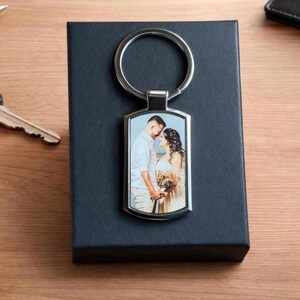 May include: A rectangular silver keychain with a photo of a couple. The keychain is on a black box. The photo shows a man and a woman embracing. The keychain has a silver ring.