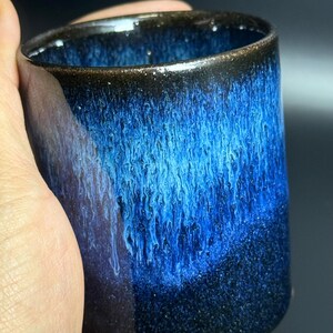 Puede incluir: Una taza de cerámica hecha a mano con un llamativo esmalte azul. La taza presenta una base azul oscuro que hace la transición a tonos de azul más claros, creando un efecto de cascada. El borde es de color marrón oscuro.