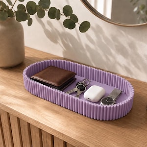 May include: An oval-shaped lavender tray with a ribbed, vertical-textured exterior wall. The tray contains a brown leather wallet, a set of metal keys, a white wireless earbud charging case, and a silver-toned wristwatch with a metal link band.