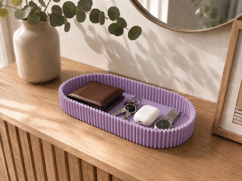 May include: An oval-shaped lavender tray with a ribbed, vertical-textured exterior wall. The tray contains a brown leather wallet, a set of metal keys, a white wireless earbud charging case, and a silver-toned wristwatch with a metal link band.