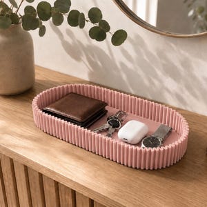 May include: An oval-shaped, dusty pink tray with a ribbed, textured exterior wall. The tray contains a dark brown leather wallet, a set of metal keys, a white wireless earbud charging case, and a silver wristwatch with a metal link band.