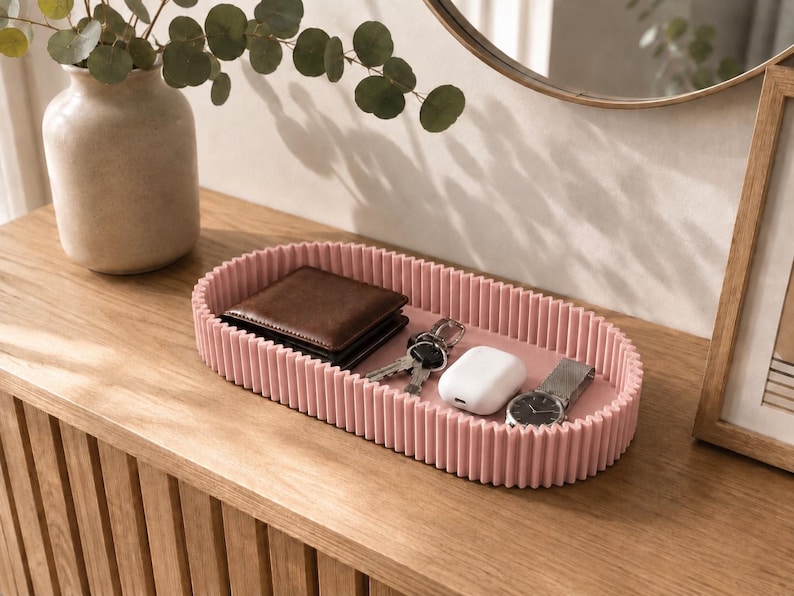 May include: An oval-shaped, dusty pink tray with a ribbed, textured exterior wall. The tray contains a dark brown leather wallet, a set of metal keys, a white wireless earbud charging case, and a silver wristwatch with a metal link band.