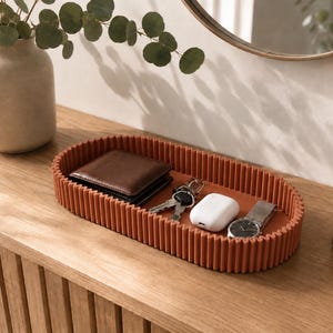 May include: An oval-shaped, terracotta-coloured tray with a ribbed, vertical-striped exterior wall. The tray contains a brown leather wallet, a set of metal keys, a white wireless earbud charging case, and a silver-toned wristwatch with a metal band.