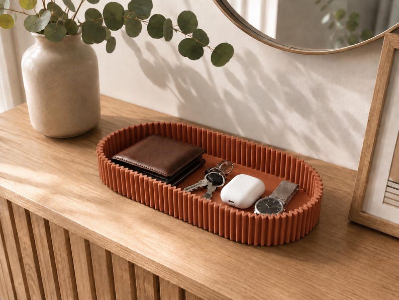 May include: An oval-shaped, terracotta-coloured tray with a ribbed, vertical-striped exterior wall. The tray contains a brown leather wallet, a set of metal keys, a white wireless earbud charging case, and a silver-toned wristwatch with a metal band.