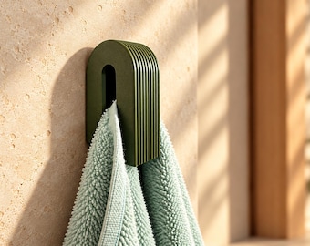 May include: A dark olive green wall-mounted hook with a rounded, arched top and vertical ribbed detailing. The hook features a central rectangular opening and is holding a light mint green textured towel with a looped fabric surface.