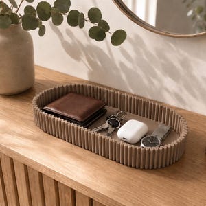 May include: An oval-shaped tray with a ribbed, taupe-coloured exterior and a smooth flat base. The tray contains a brown leather wallet, a set of metal keys, a white wireless earbud charging case, and a silver wristwatch with a metal link band.