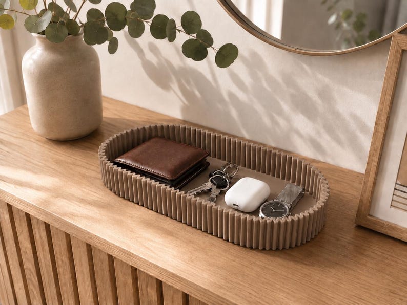 May include: An oval-shaped tray with a ribbed, taupe-coloured exterior and a smooth flat base. The tray contains a brown leather wallet, a set of metal keys, a white wireless earbud charging case, and a silver wristwatch with a metal link band.