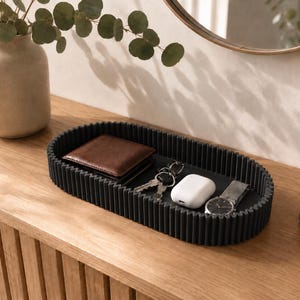 May include: An oval-shaped black tray with a ribbed, textured exterior wall. The tray contains a brown leather wallet, a set of metal keys, a white wireless earbud charging case, and a silver wristwatch with a black face and a metal link band.