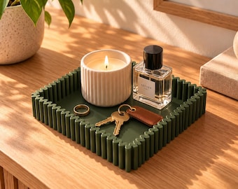 May include: A square, dark green tray with a wavy, scalloped edge. Inside the tray sits a white, ribbed candle with a lit flame, a clear glass perfume bottle with a black cap labelled AETHER, a gold-toned ring, and a set of keys with a brown tag.