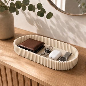 May include: An oval-shaped off-white tray with a ribbed, fluted exterior wall. The tray contains a brown leather wallet, a set of metal keys, a white wireless earbud charging case, and a silver-toned wristwatch with a dark face and metal band.