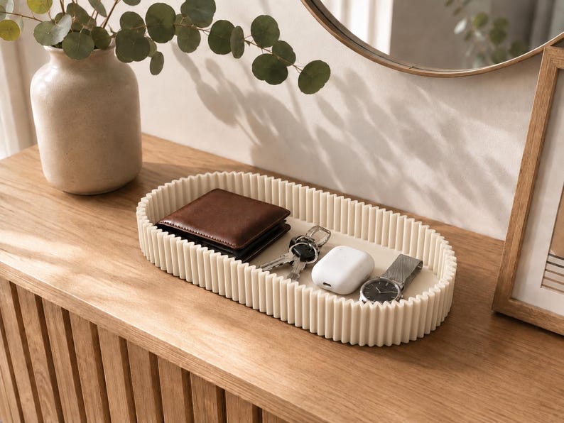 May include: An oval-shaped off-white tray with a ribbed, fluted exterior wall. The tray contains a brown leather wallet, a set of metal keys, a white wireless earbud charging case, and a silver-toned wristwatch with a dark face and metal band.
