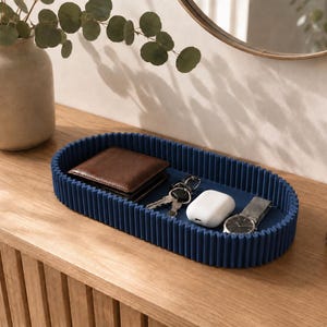 May include: An oval-shaped navy blue tray with a ribbed, fluted exterior wall. The tray contains a brown leather wallet, a set of metal keys, a white wireless earbud charging case, and a silver wristwatch with a dark face and a metal link band.