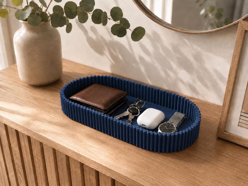 May include: An oval-shaped navy blue tray with a ribbed, fluted exterior wall. The tray contains a brown leather wallet, a set of metal keys, a white wireless earbud charging case, and a silver wristwatch with a dark face and a metal link band.