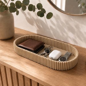 May include: An oval-shaped beige tray with a ribbed, textured exterior wall. The tray contains a brown leather wallet, a set of metal keys, a white wireless earbud charging case, and a silver-toned wristwatch with a metal link band.