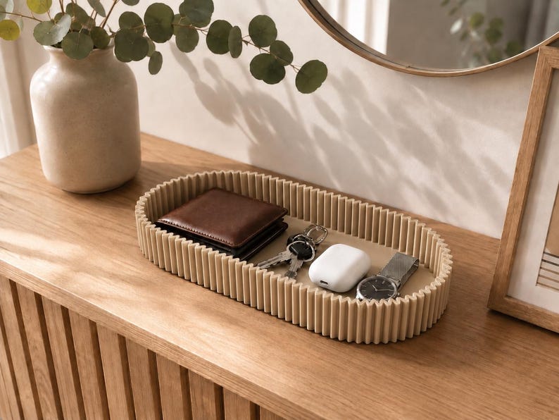 May include: An oval-shaped beige tray with a ribbed, textured exterior wall. The tray contains a brown leather wallet, a set of metal keys, a white wireless earbud charging case, and a silver-toned wristwatch with a metal link band.