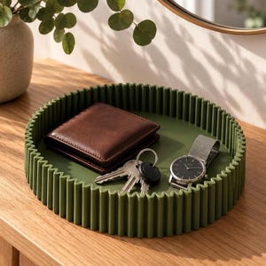 Peut inclure: Un plateau rond vert olive avec une paroi extérieure à nervures verticales. Le plateau contient un portefeuille en cuir marron, un trousseau de clés en métal sur un anneau et une montre-bracelet avec un cadran noir et un bracelet gris.