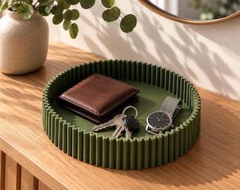 May include: A round, olive green tray with a vertically ribbed exterior wall. The tray contains a brown leather wallet, a set of metal keys on a ring, and a wristwatch with a black face and a grey strap, arranged on the flat interior base.