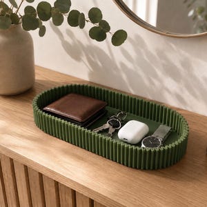 Pode incluir: Uma bandeja de armazenamento oval de cor verde escura com uma textura exterior canelada verticalmente. A bandeja contém uma carteira de couro castanha, um conjunto de chaves de metal, um estojo de carregamento branco e um relógio de pulso prateado.