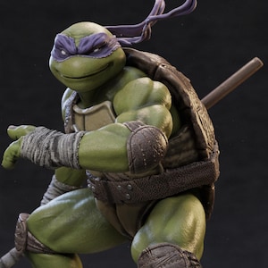 May include: A detailed figurine of Donatello from Teenage Mutant Ninja Turtles. The figure is predominantly green with a purple mask, brown shell, and arm wraps. He is holding a bo staff, showcasing a dynamic pose.