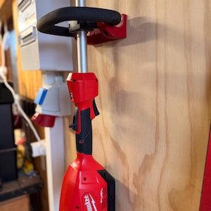 Op de afbeelding: Een rood-zwarte Milwaukee FUEL-trimmer gemonteerd op een houten muur. De trimmer wordt op zijn plaats gehouden door een rode beugel. De handgreep is zwart en gebogen. De behuizing van de trimmer is rood met zwarte accenten en het Milwaukee-logo.