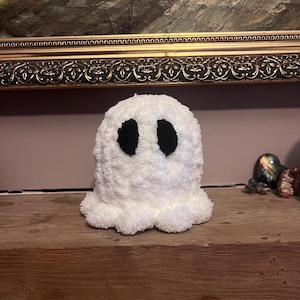 May include: A fluffy white ghost-shaped plush toy with two large black eyes. The soft toy is made from a textured material and is displayed on a wooden surface. A decorative item, perfect for Halloween.