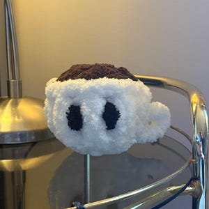 May include: A plush, crocheted coffee mug toy made of soft white yarn with a dark brown textured top representing coffee. The mug features two round black yarn eyes on the front and a small white handle on the side, crafted in a rounded shape.