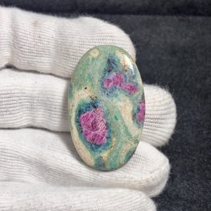 Puede incluir: Un cabujón de piedra preciosa pulida de forma ovalada con una superficie moteada en tonos verde claro, crema y azul oscuro, acentuado por tres inclusiones cristalinas de color magenta vibrante incrustadas en la matriz de la piedra.