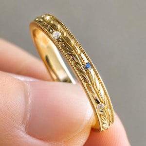 Puede incluir: Un anillo de oro amarillo con un patrón de hojas grabado en la superficie exterior. El anillo está decorado con dos pequeñas piedras redondas transparentes y una pequeña piedra redonda azul engastadas a intervalos en el diseño de hojas.