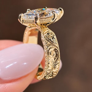 May include: A gold ring featuring a large marquise-cut clear gemstone held by multiple prongs. The band is crafted from yellow gold and displays intricate floral scrollwork engravings along the sides, accented by a fine milgrain beaded edge.