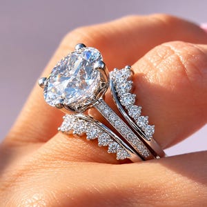 May include: A silver-toned ring set featuring a large oval-cut center stone with a prong setting, flanked by two curved bands adorned with multiple small round-cut stones. The central band has a pavé-set shank, creating a layered, sparkling appearance.