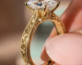 May include: A gold ring featuring a large, rectangular, faceted clear gemstone set in a four-prong mounting. The band is crafted from yellow gold and displays intricate, engraved scrollwork patterns along the entire outer surface of the shank.
