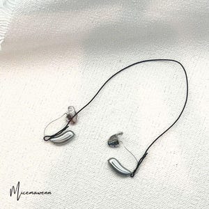Può includere: Due piccoli apparecchi acustici color argento con fili neri e auricolari trasparenti sono esposti su una superficie bianca. Gli apparecchi acustici hanno una forma oblunga e curva. Gli auricolari trasparenti sono visibili e l'estetica generale è pulita e moderna.