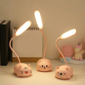 May include: Three pink cat-shaped desk lamps with flexible necks and bright, rectangular LED lights. Each lamp has a different facial expression. The lamps are on a white surface.