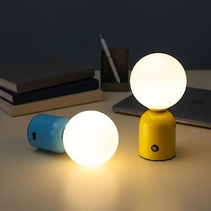 May include: Two small, modern lamps with a spherical light. One lamp is yellow, the other is blue. Both have a round, glowing light. The yellow lamp is upright, while the blue lamp is lying on its side. The lamps are on a light-colored surface.