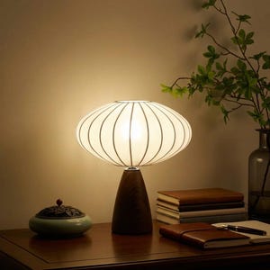 Peut inclure: Une lampe de table moderne avec un abat-jour blanc de forme ovale orné de lignes noires verticales. La lampe repose sur un socle en bois conique marron foncé. Sur une table en bois, des livres, un carnet et un brûleur d'encens décoratif.