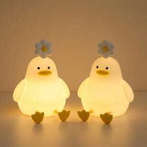 May include: Two white duck-shaped night lights with yellow beaks and feet. Each duck has a white flower on its head. The lights emit a soft, warm glow, creating a cosy ambiance. The background is a neutral colour.