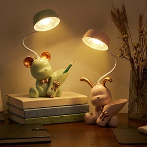 Può includere: Due lampade da scrivania fantasiose, una a forma di orso verde e l'altra di coniglio rosa, ciascuna con un portapenne. Le lampade hanno colli bianchi regolabili e paralumi rotondi. Una matita è nel portapenne dell'orso.
